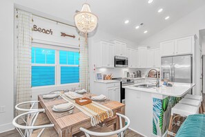 A modern kitchen with white cabinetry, a dining area with a wooden table and white chairs, a chandelier, and a view of the beach through a window.