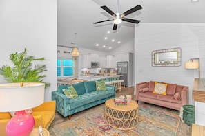 A modern living room with a teal sofa, a pink sofa, and a coffee table. There is a kitchen in the background with white cabinets and a ceiling fan. - Beach Cottage: Glow Arcade + Aquarium Bunk Room (Myrtle Beach)