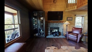 A cozy cabin interior with a stone fireplace, leather armchair, and a large TV mounted on the wall. - Amazing Views!! Creekside Cabin, Wrap Around Porch, Close to Town! Dogs Welcome! (Hiawassee)