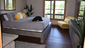 A bedroom with a large bed, a wooden headboard, a window with a view of greenery, a yellow armchair, and a potted plant. - Amazing Views!! Creekside Cabin, Wrap Around Porch, Close to Town! Dogs Welcome! (Hiawassee)