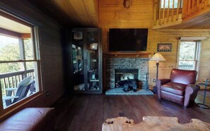 A cozy cabin interior with a stone fireplace, leather armchair, and a large TV mounted on the wall. - Amazing Views!! Creekside Cabin, Wrap Around Porch, Close to Town! Dogs Welcome! (Hiawassee)