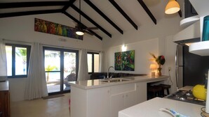 A modern kitchen with a central island, a dining area with a sofa, and a large window offering a view of the outdoors. - Tropical 1 Bedroom Cabana on Private Oasis (Maya Beach)