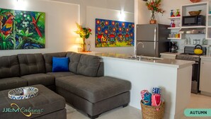 A modern kitchen with a grey sofa, a dining table, and a colorful painting above the counter. - Tropical 1 Bedroom Cabana on Private Oasis (Maya Beach)