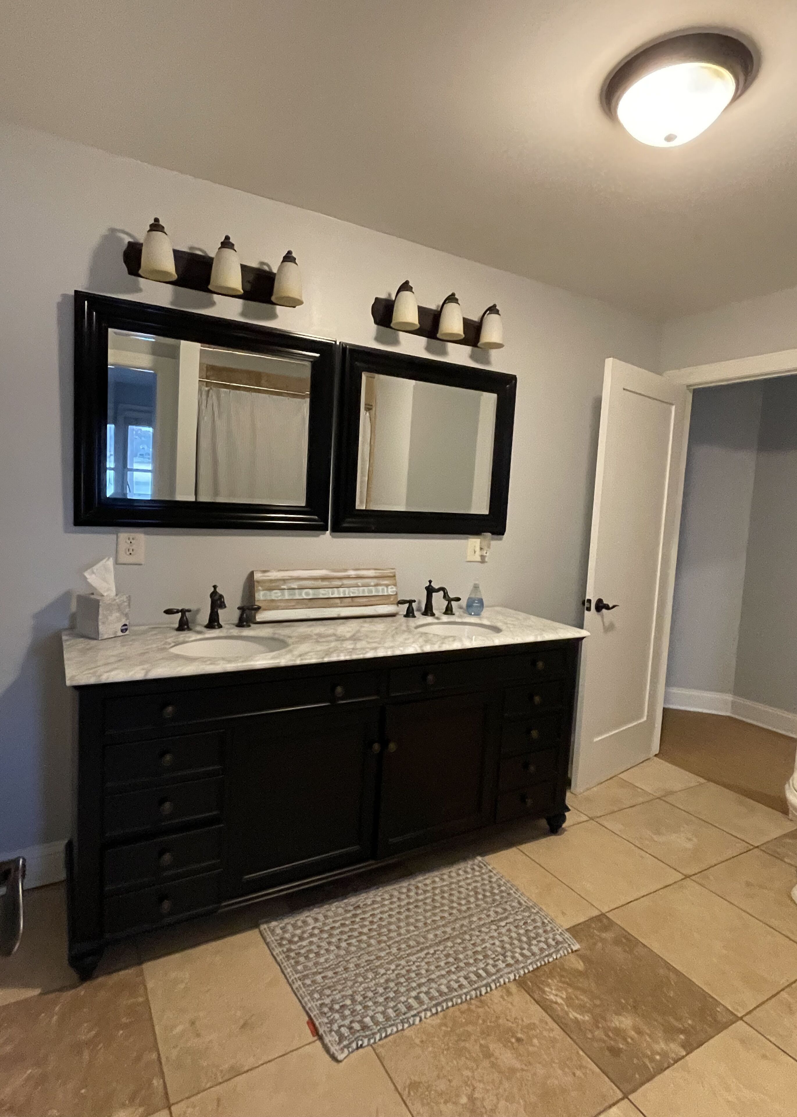 Un bagno con doppio lavabo, due specchi, un lavandino bianco, mobili scuri, un portasciuga e un lampadario sopra gli specchi.