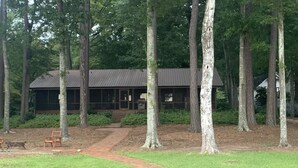 Un chalet doté d’une grande façade vitrée, entouré d’une zone boisée et d’une jetée s’avançant dans un plan d’eau.