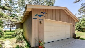 A wooden garage with a garage door, surrounded by greenery and a clear sky.