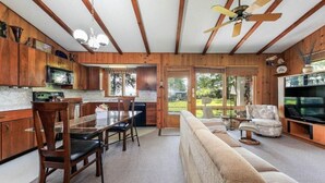 A cozy cabin interior with wooden walls, a dining area, a living room with a sofa and TV, and a kitchen with a microwave.