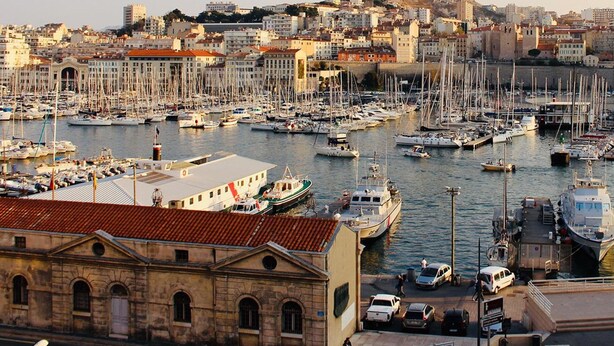 Una marina con numerose imbarcazioni ormeggiate, un edificio storico con tetto in tegole rosse e uno skyline sullo sfondo.