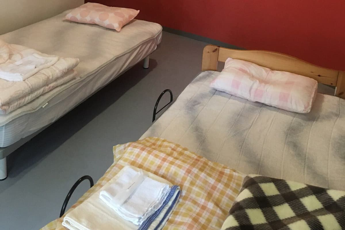 Een kamer met twee enkele bedden, elk met een kussen en een dekbed. Er staat een klein tafeltje met een opgevouwen handdoek en een geruit tafelkleed. De muren zijn rood en de vloer is grijs.