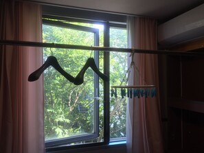 A window with a view of trees, curtains, and a clothing rack with items hanging. - Narita Airport Samurai Hostel (Kujukuri)