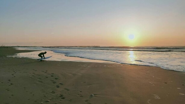 Une plage où l’on voit des empreintes de pas menant à l’eau, une personne près du bord de l’eau et le soleil se couchant à l’horizon.