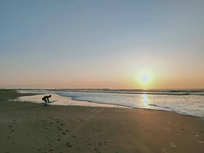 A beach with footprints leading to the water, a person near the water's edge, and the sun setting over the horizon.