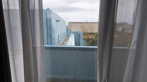 Une vue à travers une fenêtre ornée de rideaux légers, dévoilant un balcon bleu avec une rambarde en métal, un sol en béton et un bâtiment en arrière-plan.