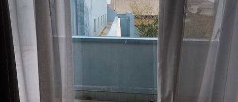 Uitzicht door een raam met dunne gordijnen, waarop een blauw balkon met metalen hekwerk, een betonnen vloer en een gebouw op de achtergrond zichtbaar is.