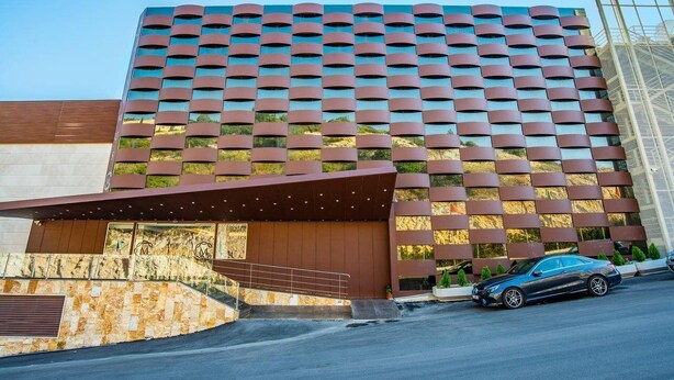 A modern hotel with a distinctive facade featuring a series of horizontal metallic panels and a large entrance with a covered walkway. - Maximus Hotel (Blat)