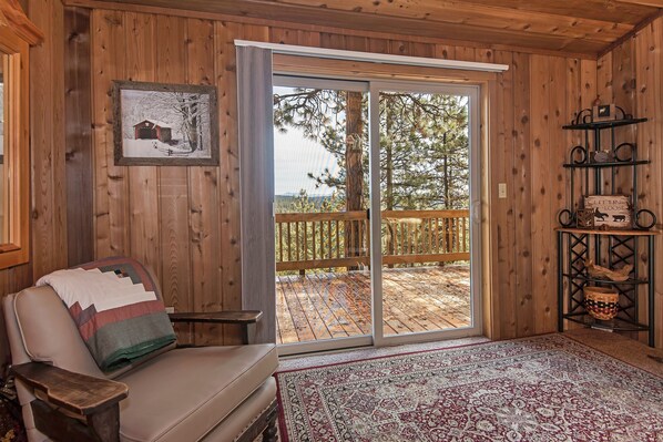 A cozy cabin interior with a sliding glass door leading to a deck, a cushioned chair, a patterned rug, and a shelf with decorative items.