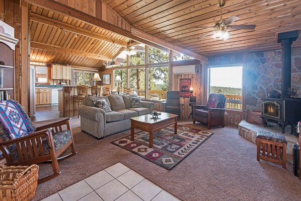 A spacious living area with a fireplace, wooden furniture, and a large window overlooking a forested area.