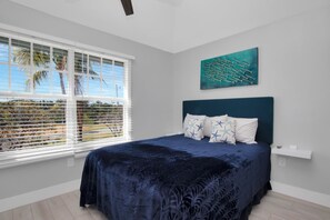 A bedroom with a bed, a window with blinds, a wall-mounted shelf, and a painting.