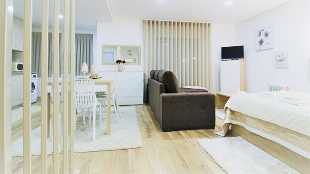A modern hotel room with a wooden floor, a bed, a sofa, a dining table with chairs, a TV, and a radiator. - Imperial Apartment (Braga)