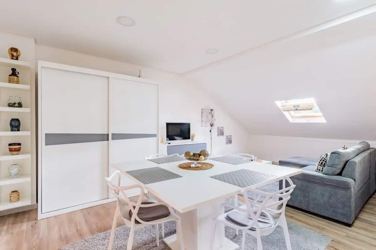 A modern, well-lit room with a dining table, chairs, a grey sofa, and a TV.