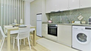 A modern kitchen with white cabinets, a granite countertop, a built-in oven, a washing machine, and a dining area with a table and chairs. - Imperial Apartment (Braga)