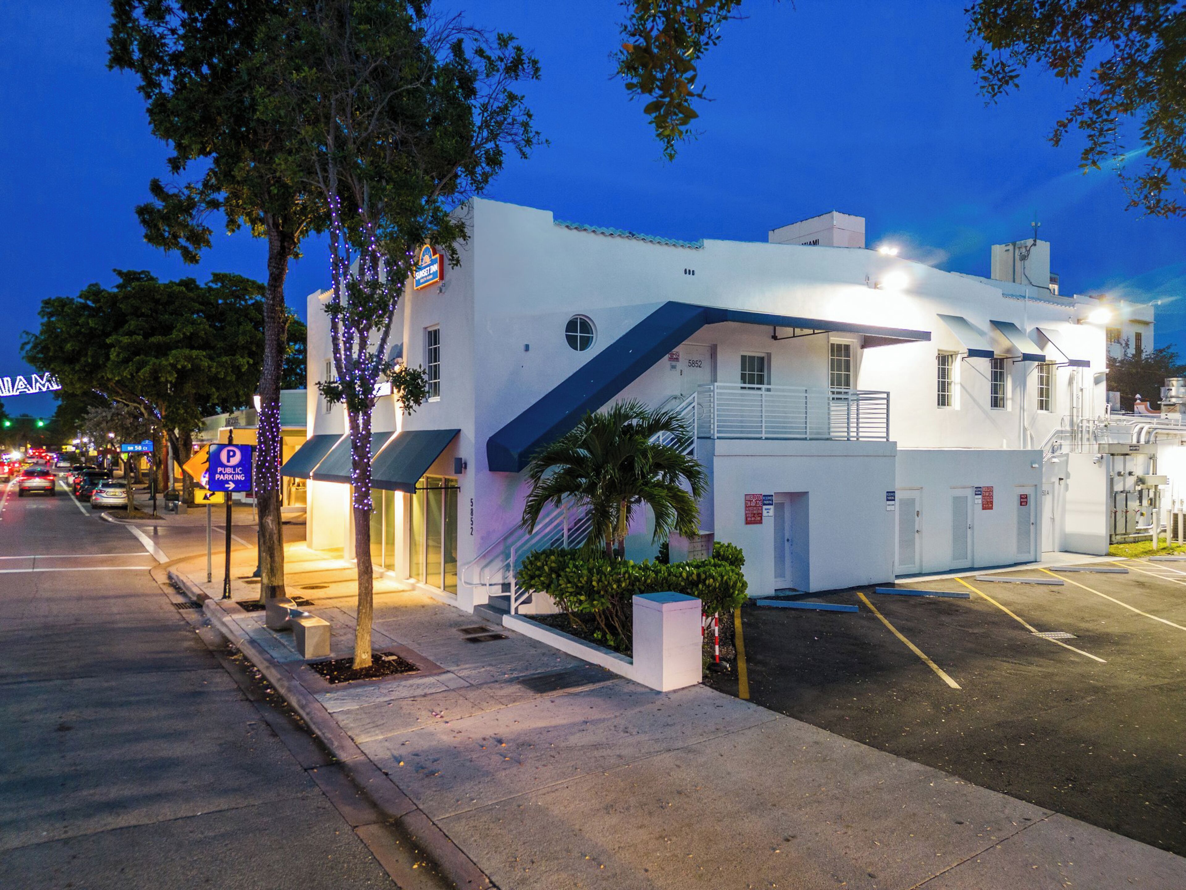 A white building with a blue awning, a parking lot, and a street with traffic.