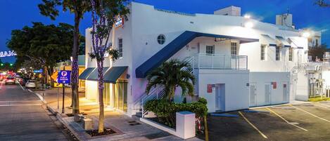 A white building with a blue awning, a parking lot, and a street with traffic.