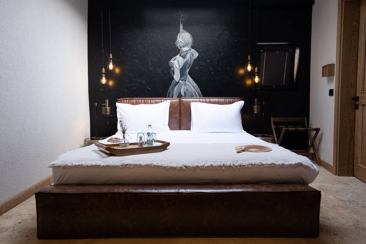 Un dormitorio con una cama grande, un banco de madera, una bandeja con vasos y una botella, y un cuadro de una mujer en la pared.
