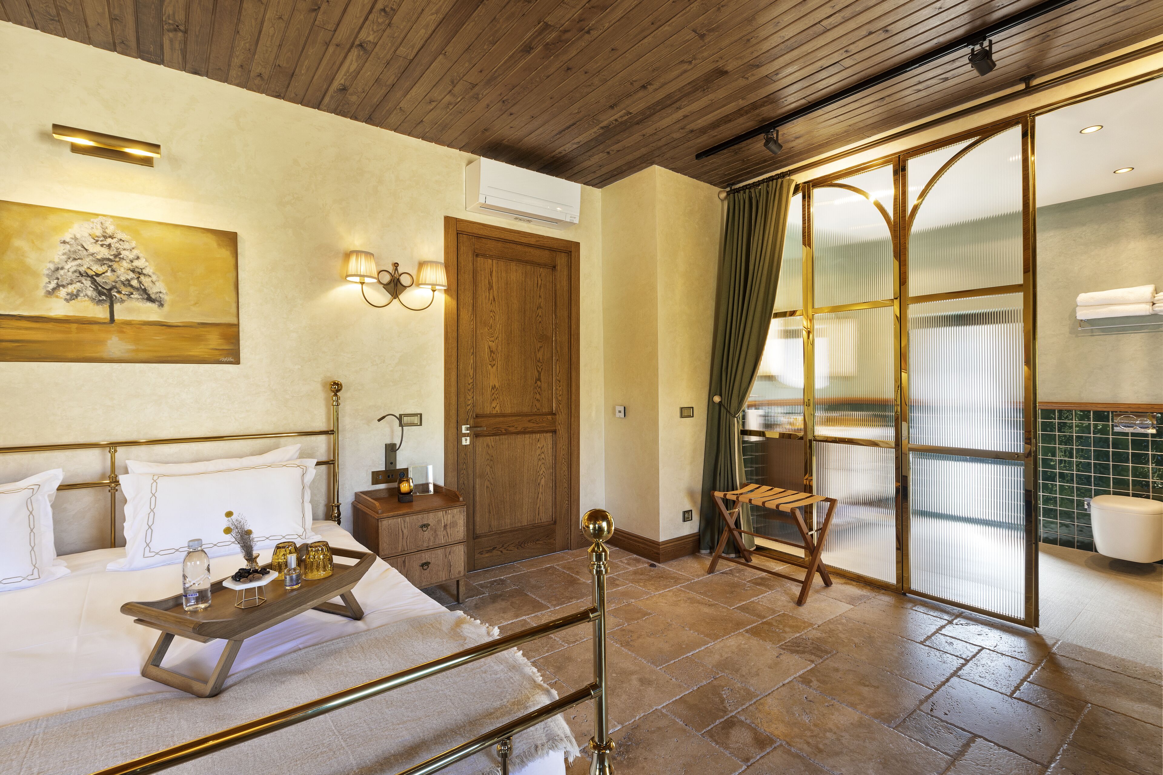 Habitación de hotel con una cama, una mesita de noche de madera, una bandeja con objetos, un cuadro, una puerta y un baño visible a través de un panel de vidrio.