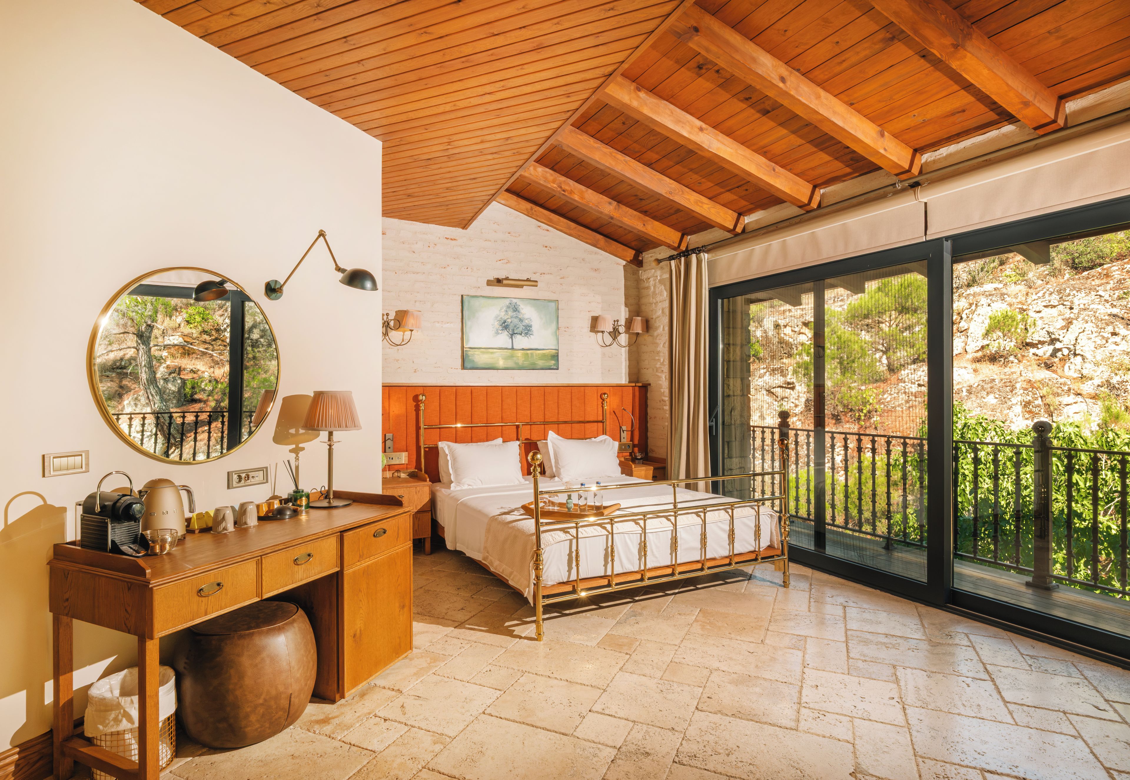 A bedroom with a wooden ceiling, a bed with a metal frame, a desk with a mirror, and a view of greenery through a large window.