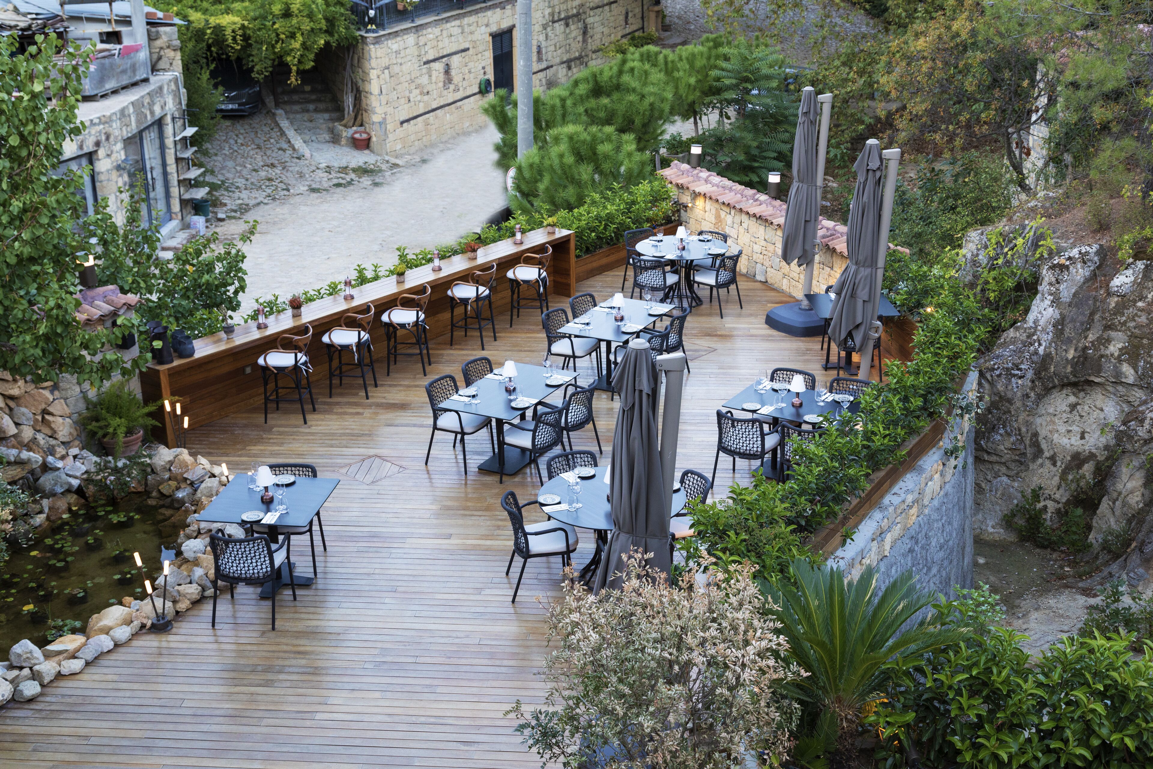 An outdoor patio with multiple tables and chairs, surrounded by greenery and a small water feature.