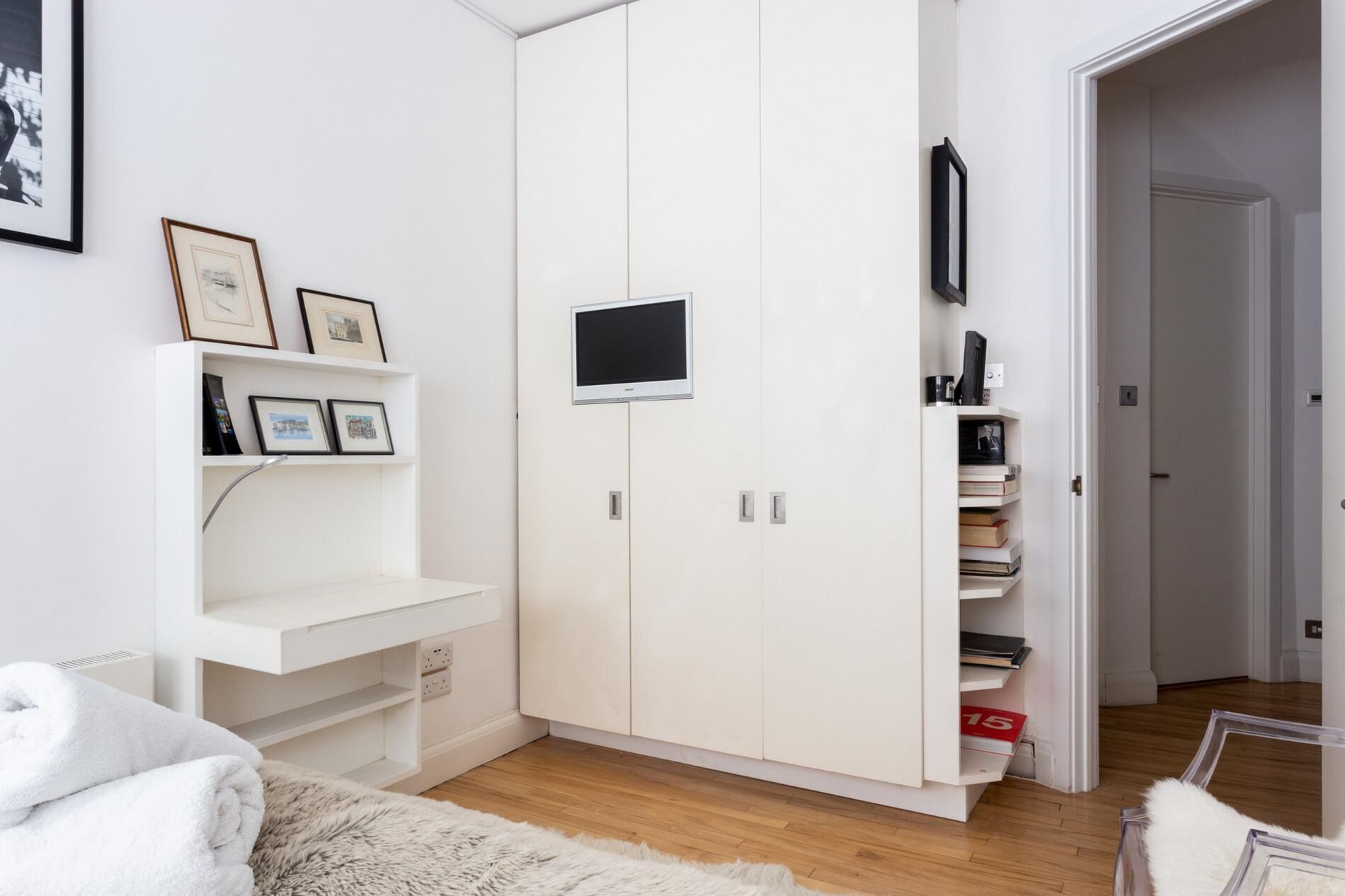 A room with a white wardrobe, a flat-screen TV mounted on the wall, and a bookshelf with framed pictures.