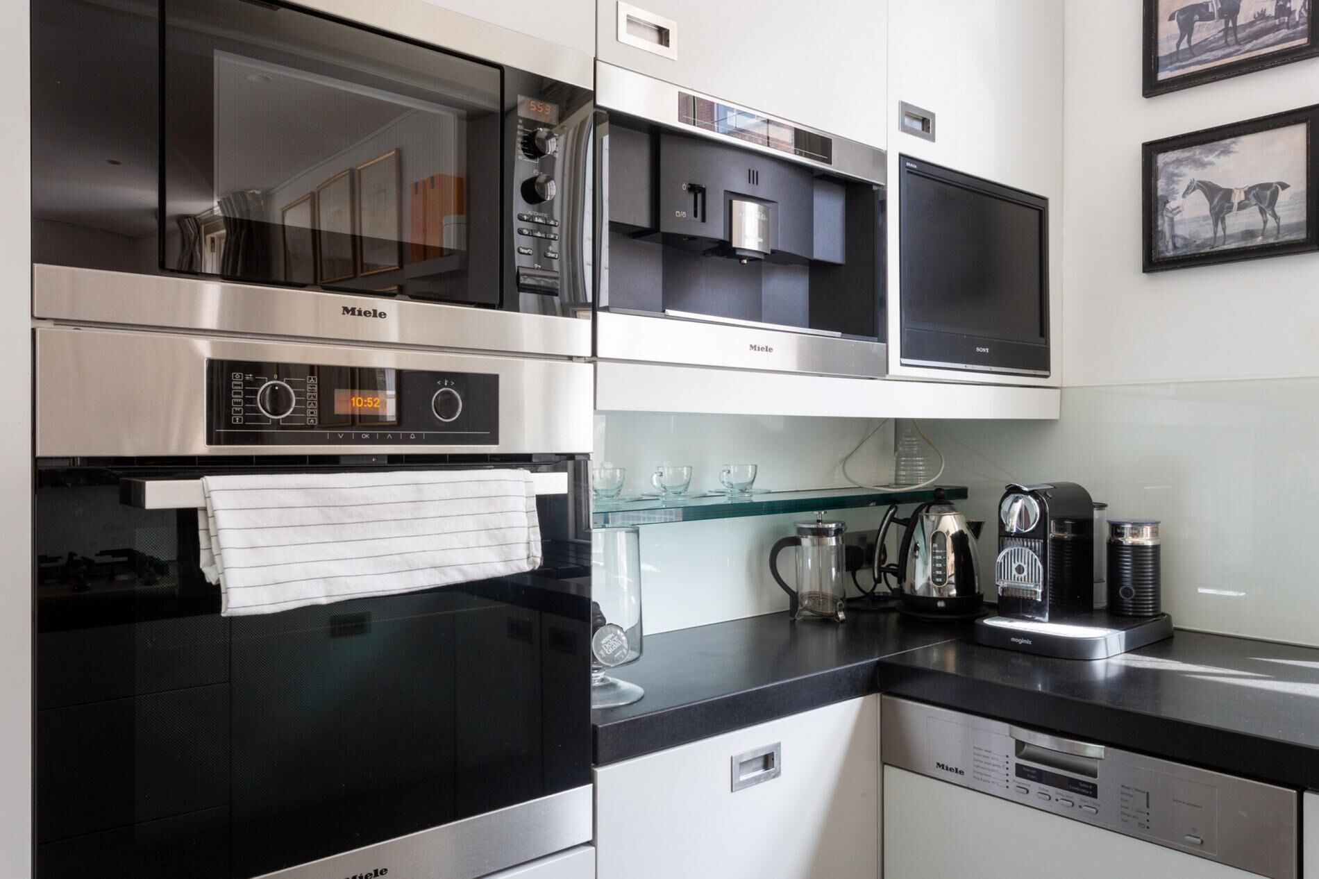 Una cocina moderna con electrodomésticos de acero inoxidable, incluyendo estufa, horno y microondas. Hay una repisa de vidrio con tazas y una cafetera.