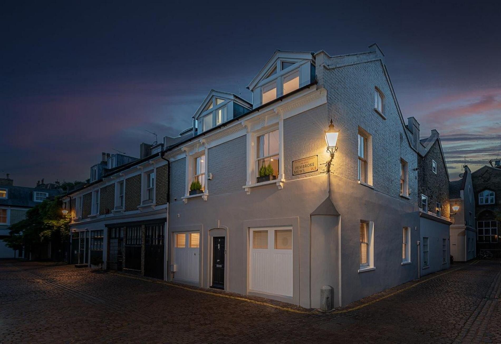 Ein traditionelles Gebäude mit einem Schild, auf dem "Pembroke Mews" steht. Es hat einen Hauseingang an der Ecke mit einer Tür, ein kleines Fenster mit einer Pflanze und eine Straßenlaterne.