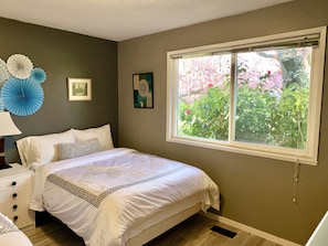 A bedroom with a bed, a nightstand, a dresser, a window with a view of greenery, and wall decorations.
