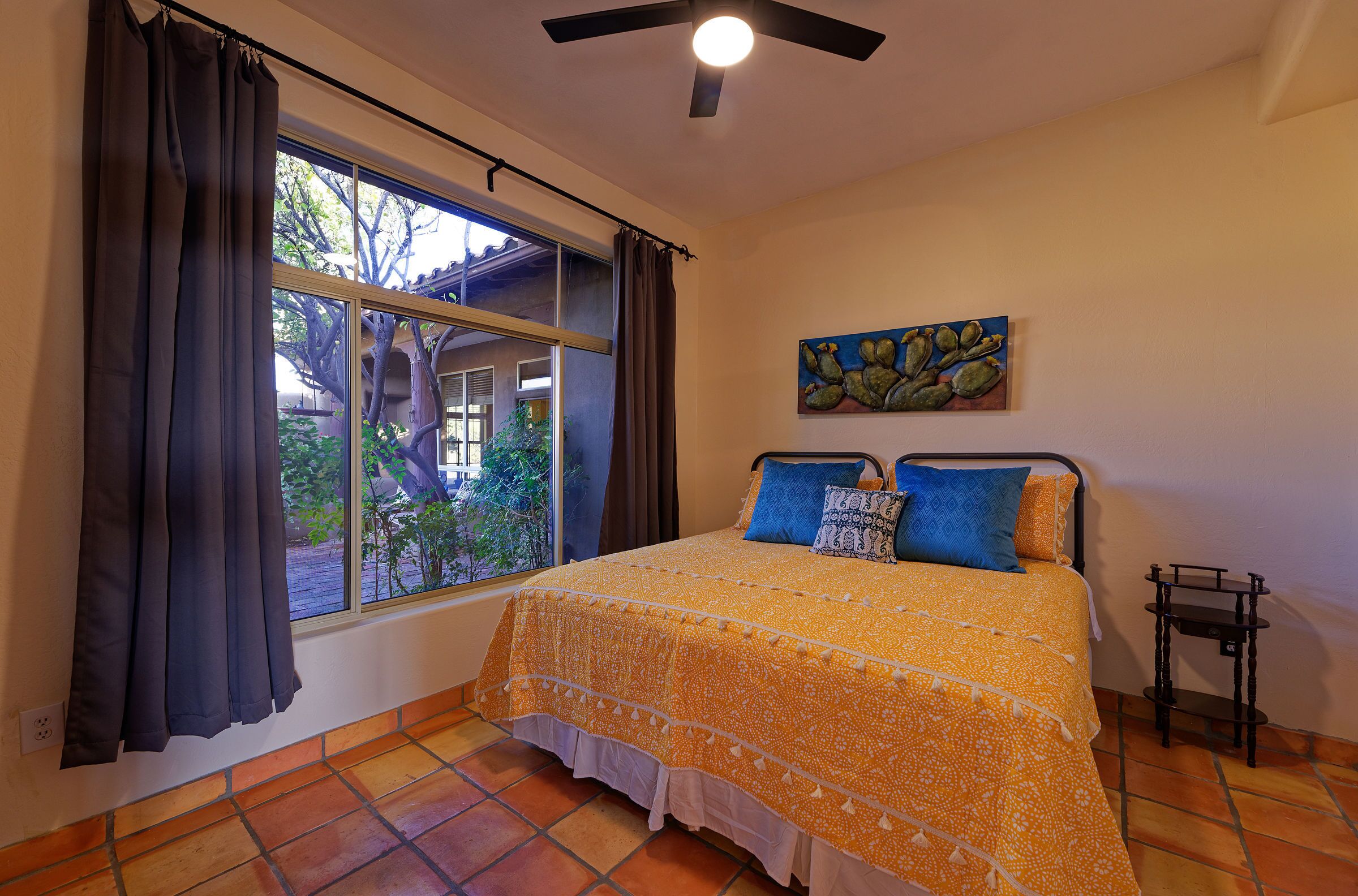 A bedroom with a bed, curtains, a ceiling fan, a window with a view, and a painting on the wall.