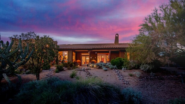 Une maison de style désertique, avec un toit en tuiles, entourée de cactus et d’arbustes, sous un ciel crépusculaire.