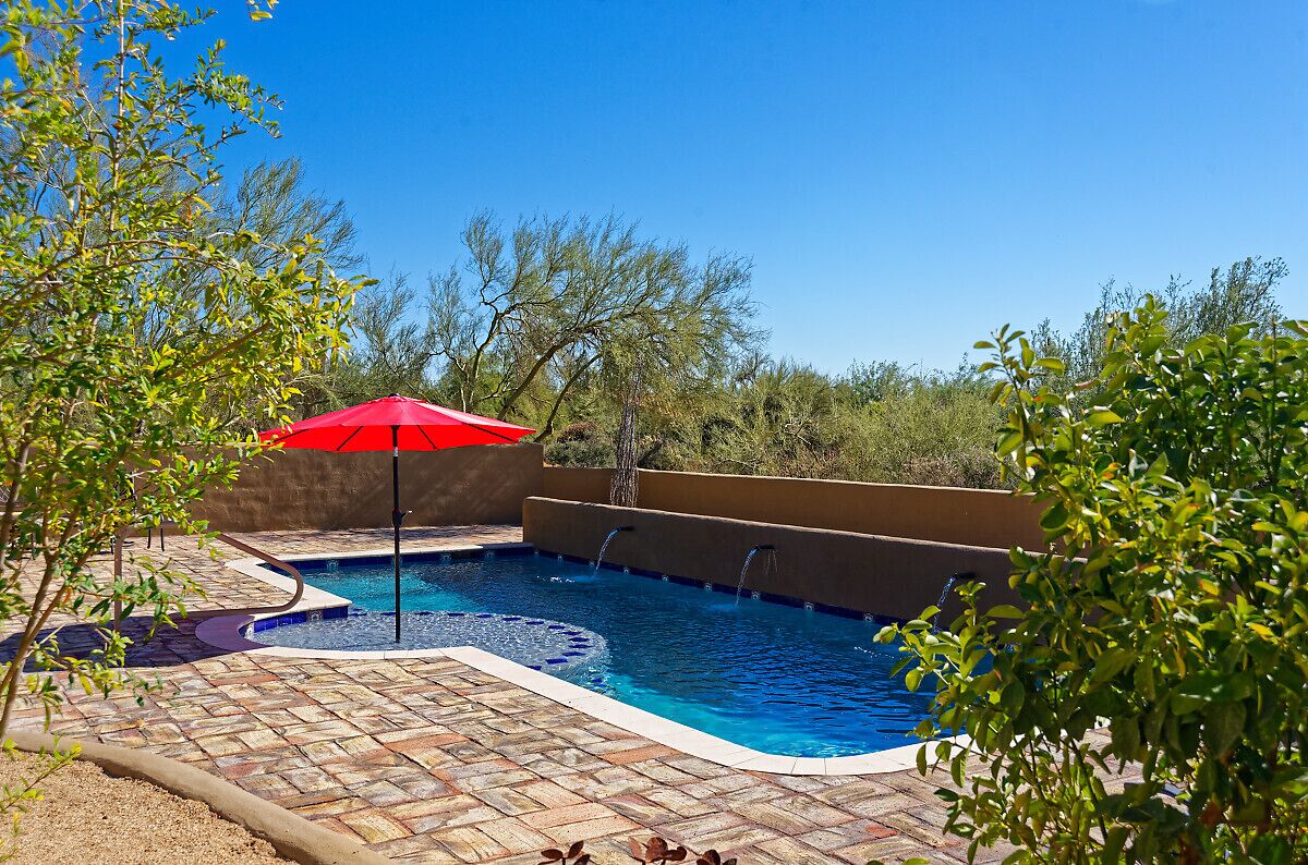 A swimming pool with a red umbrella, surrounded by a brick patio and greenery.