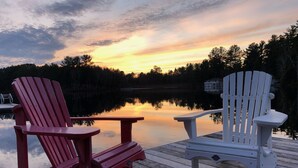 Zwei Adirondack-Stühle auf einem hölzernen Steg mit Blick auf einen stillen See, im Hintergrund ein Sonnenuntergang.