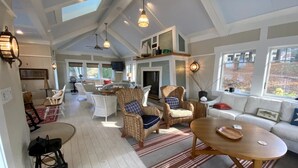 Ein geräumiger Wohnbereich mit hoher Decke, großen Fenstern und Kamin. Es gibt Korbstühle, ein Sofa und einen Esstisch.
