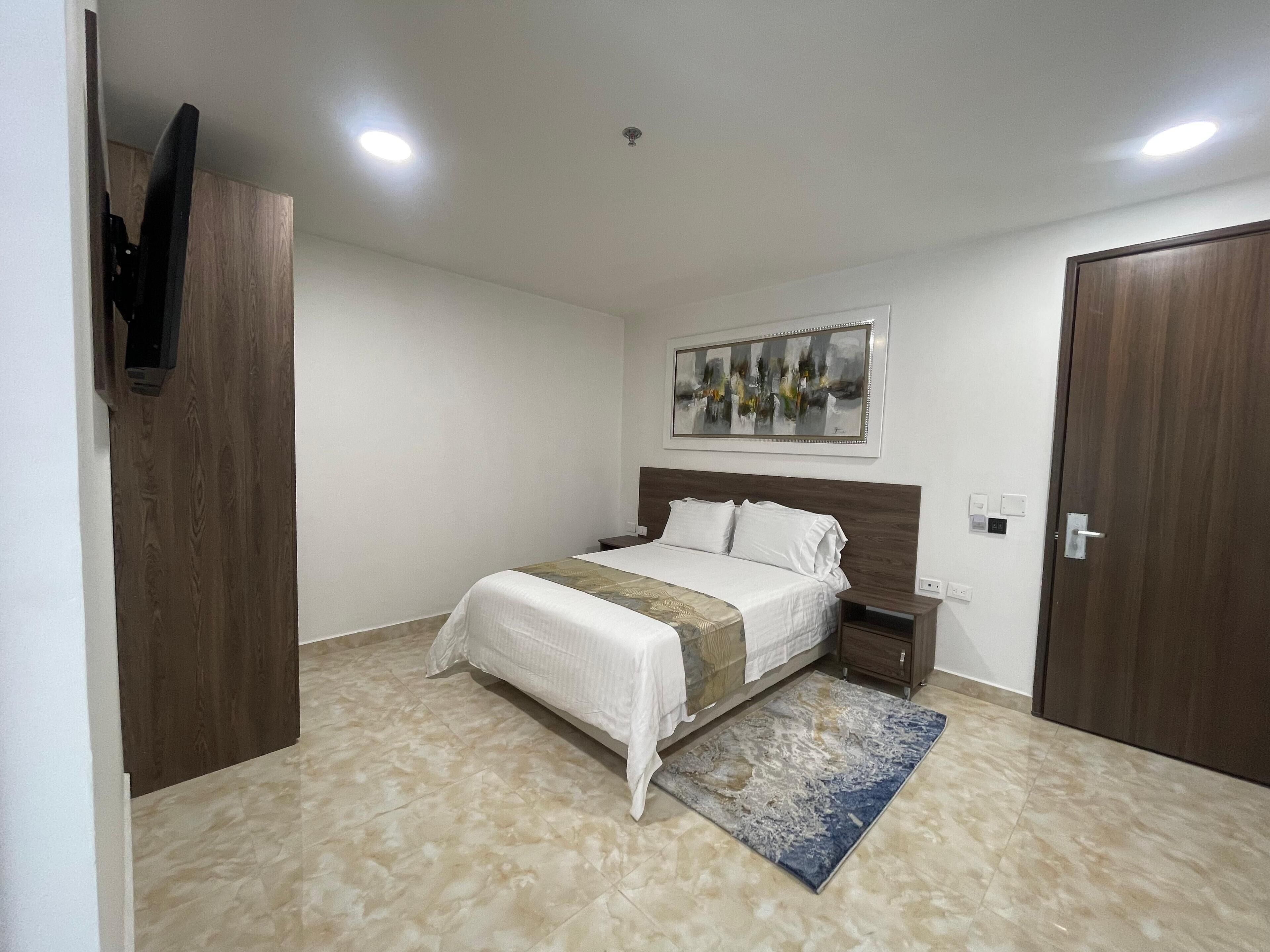 A bedroom with a bed, a nightstand, a TV mounted on a wooden door, a painting on the wall, and a door with a handle.