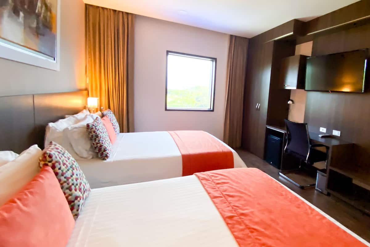 A hotel room with two beds, a desk, a chair, a TV, and a window with curtains.