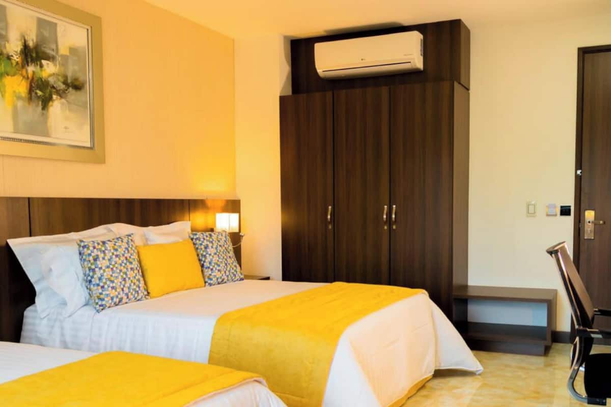A hotel room with two beds, a desk, a chair, an air conditioning unit, and a door.