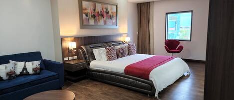A hotel room with a bed, a blue sofa, a red armchair, a wooden nightstand with lamps, and a large window with a view.