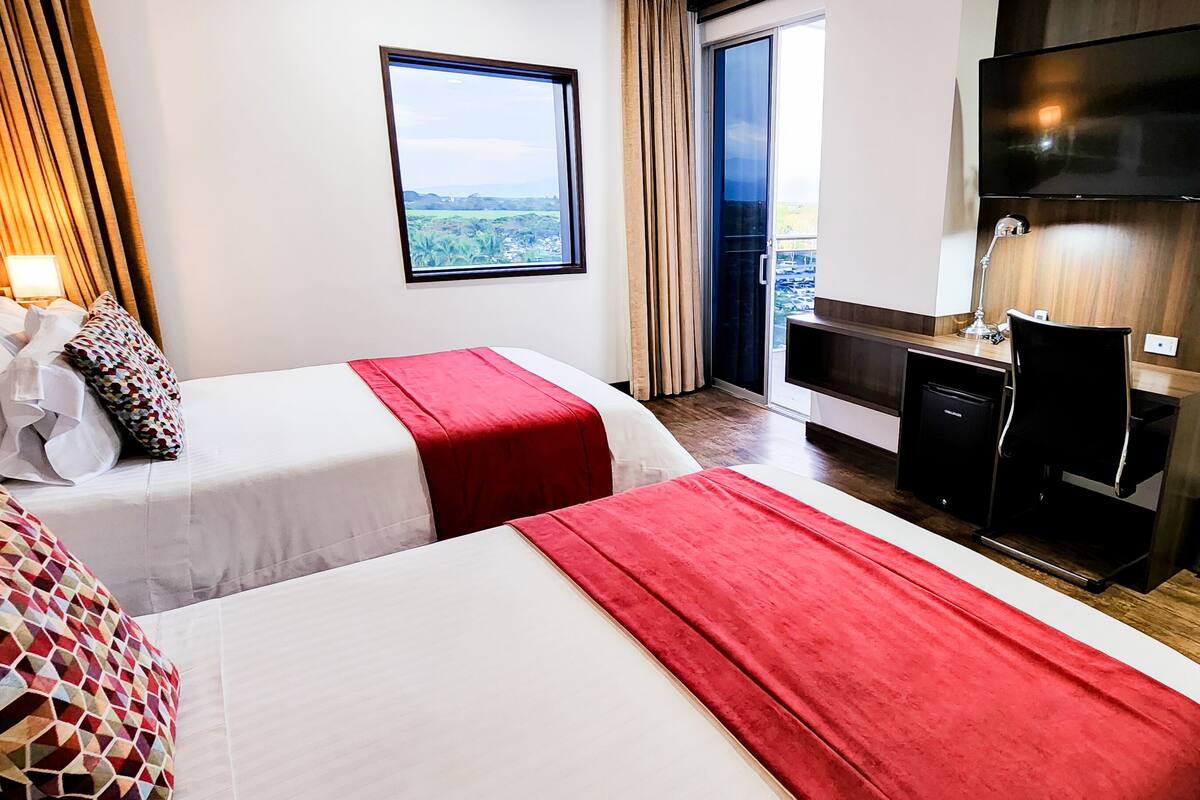 A hotel room with two beds, a desk, a TV, and a large window offering a view.