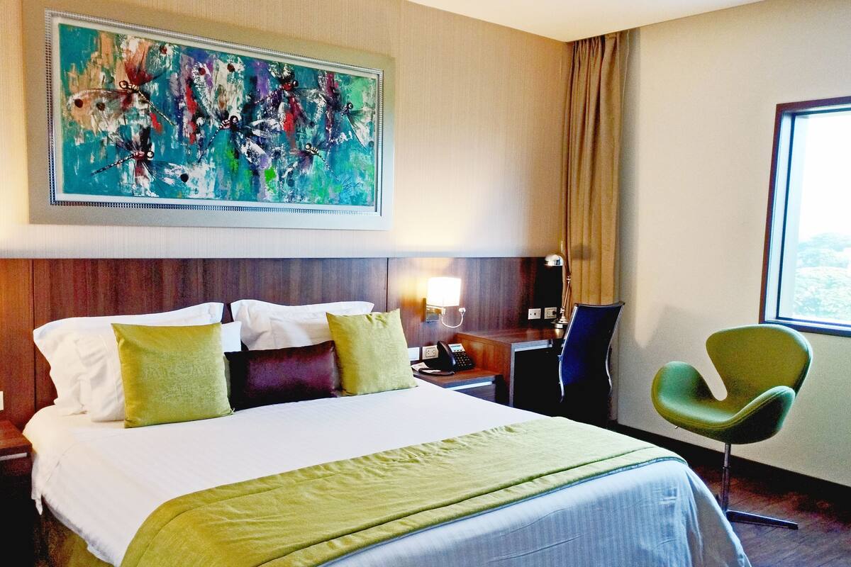 A hotel room with a large bed, a desk with a lamp, a green chair, and a painting on the wall.