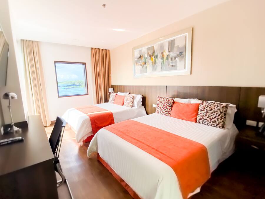 Habitación de hotel con dos camas, un escritorio, una silla, un ventanal con vistas y un cuadro en la pared.