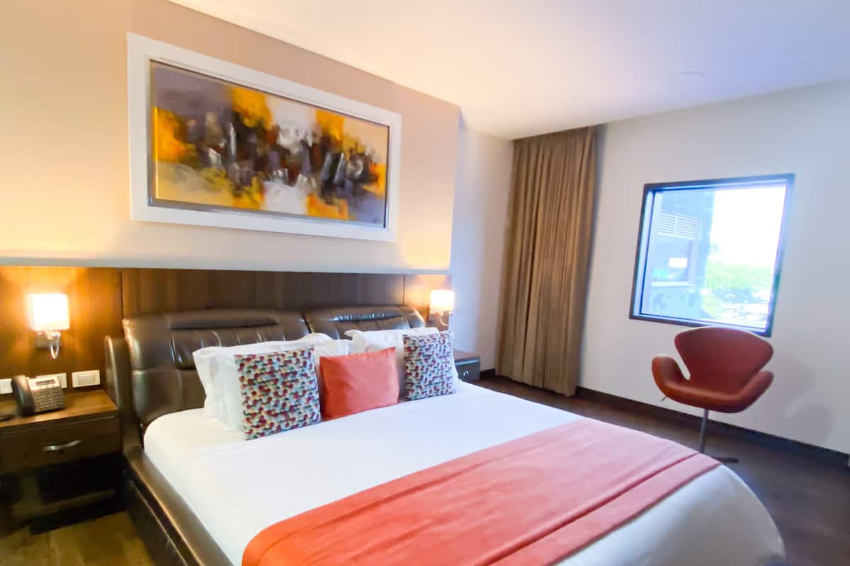 A hotel room with a large bed, a headboard, a bedside table with a phone, a red chair, a window, and a painting on the wall.