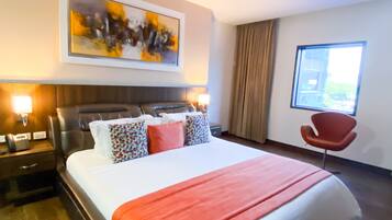 A hotel room with a large bed, a headboard, a bedside table with a phone, a red chair, a window, and a painting on the wall.