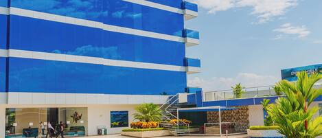 A modern hotel with a large pool area, a well-maintained garden, and a clear blue sky.